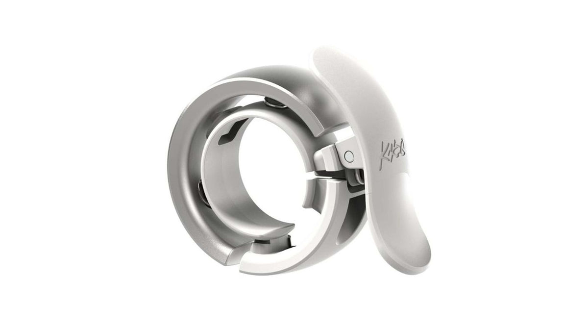 Die metallische, kreisrunde Knog OI Prima Fahrradklingel von KNOG mit klappbarem, geschwungenem Arm streckt sich nach außen - ihre glatte, silberne Oberfläche auf weißem Grund ist Ausdruck von präzisem, CNC-gefrästem Aluminium und raffiniertem Design.
