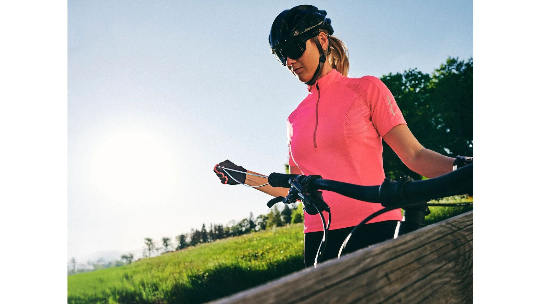 Eine Frau im rosa Fahrradtrikot, mit Helm und Sonnenbrille steht im Freien neben ihrem Fahrrad, das mit dem DIVERSE Lenklock Fahrradschloss 130cm gesichert ist, und hält den Lenker vor einer Kulisse aus grünem Gras und Bäumen unter einem strahlenden Himmel.