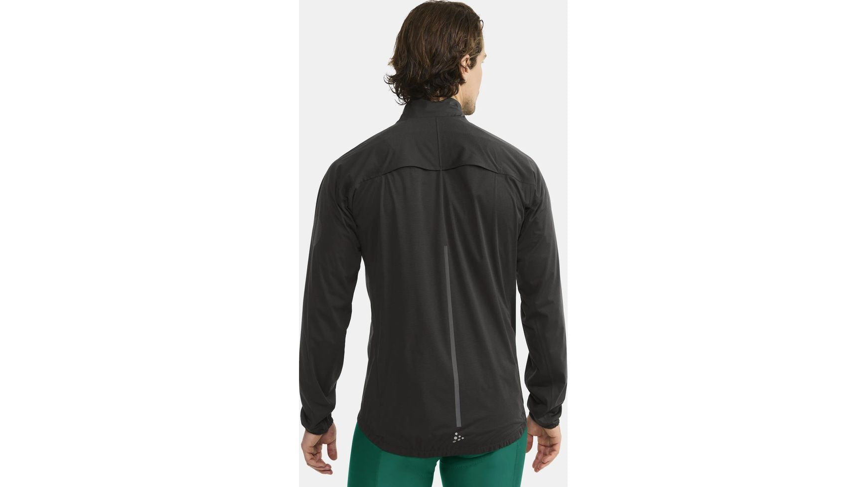 Gezeigt wird eine Person mit mittellangen braunen Haaren von hinten, die eine CRAFT ADV Hydro Jacket 2 M in Schwarz und eine grüne Hose trägt und vor einem schlichten hellen Hintergrund steht.
